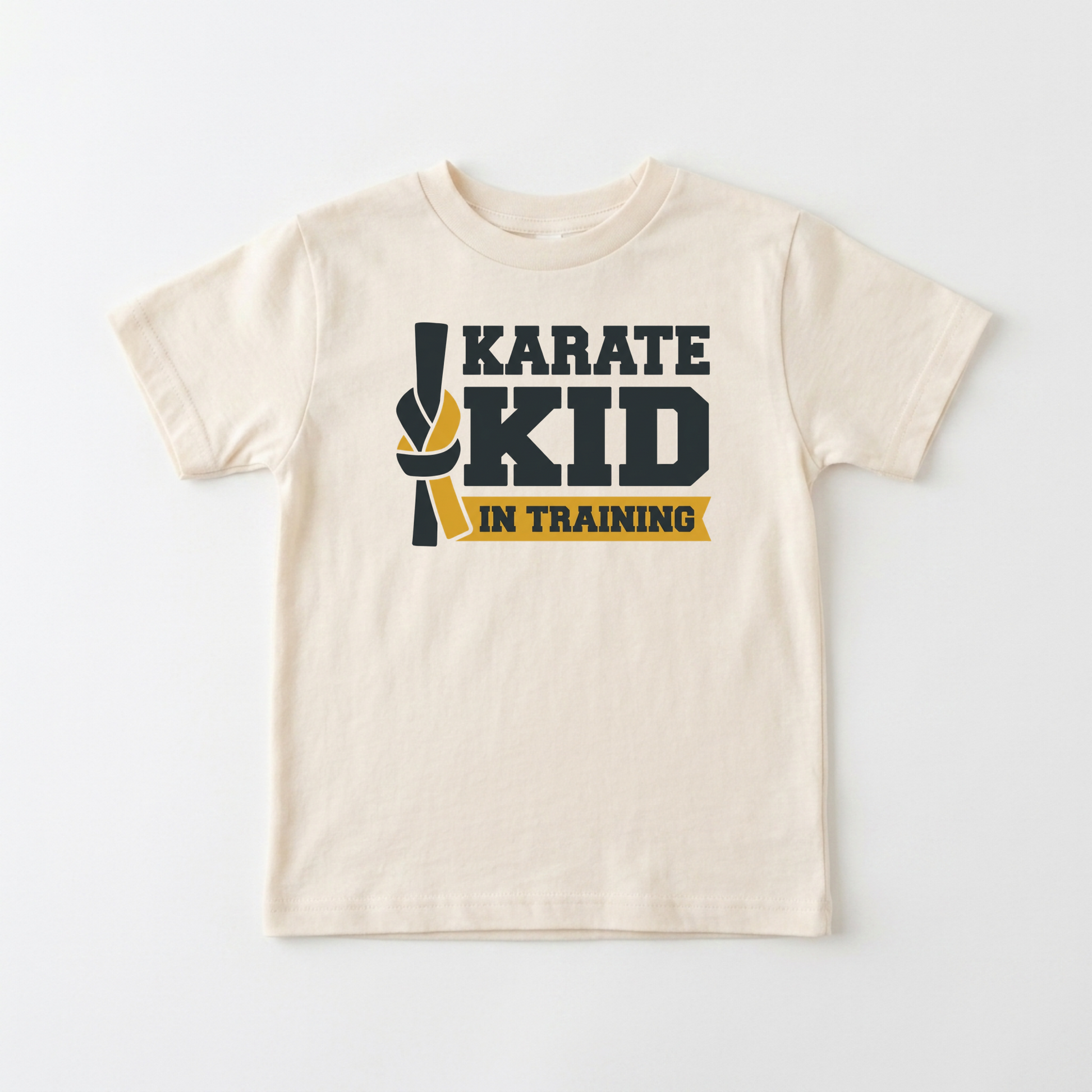 A cream-colored short-sleeve kids' t-shirt with a 'KARATE KID IN TRAINING' graphic on the chest, featuring a black and yellow martial arts ribbon design, laid flat against a light gray background.
