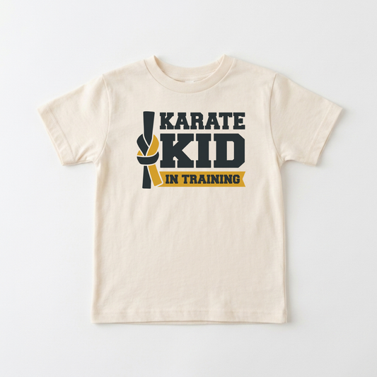 A cream-colored short-sleeve kids' t-shirt with a 'KARATE KID IN TRAINING' graphic on the chest, featuring a black and yellow martial arts ribbon design, laid flat against a light gray background.