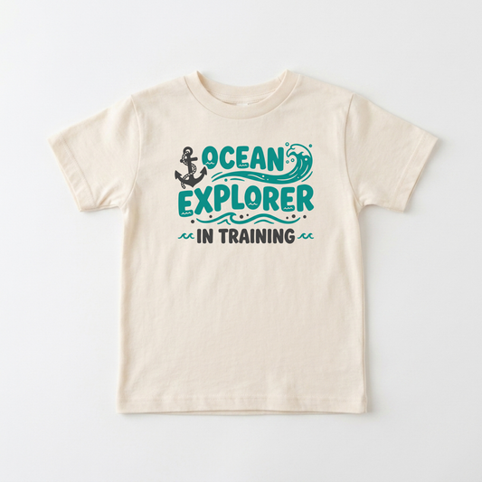 A cream-colored short-sleeve t-shirt with a graphic on the front that reads 'OCEAN EXPLORER IN TRAINING' in teal and dark gray, featuring an anchor and wave illustration, shown against a plain white background.