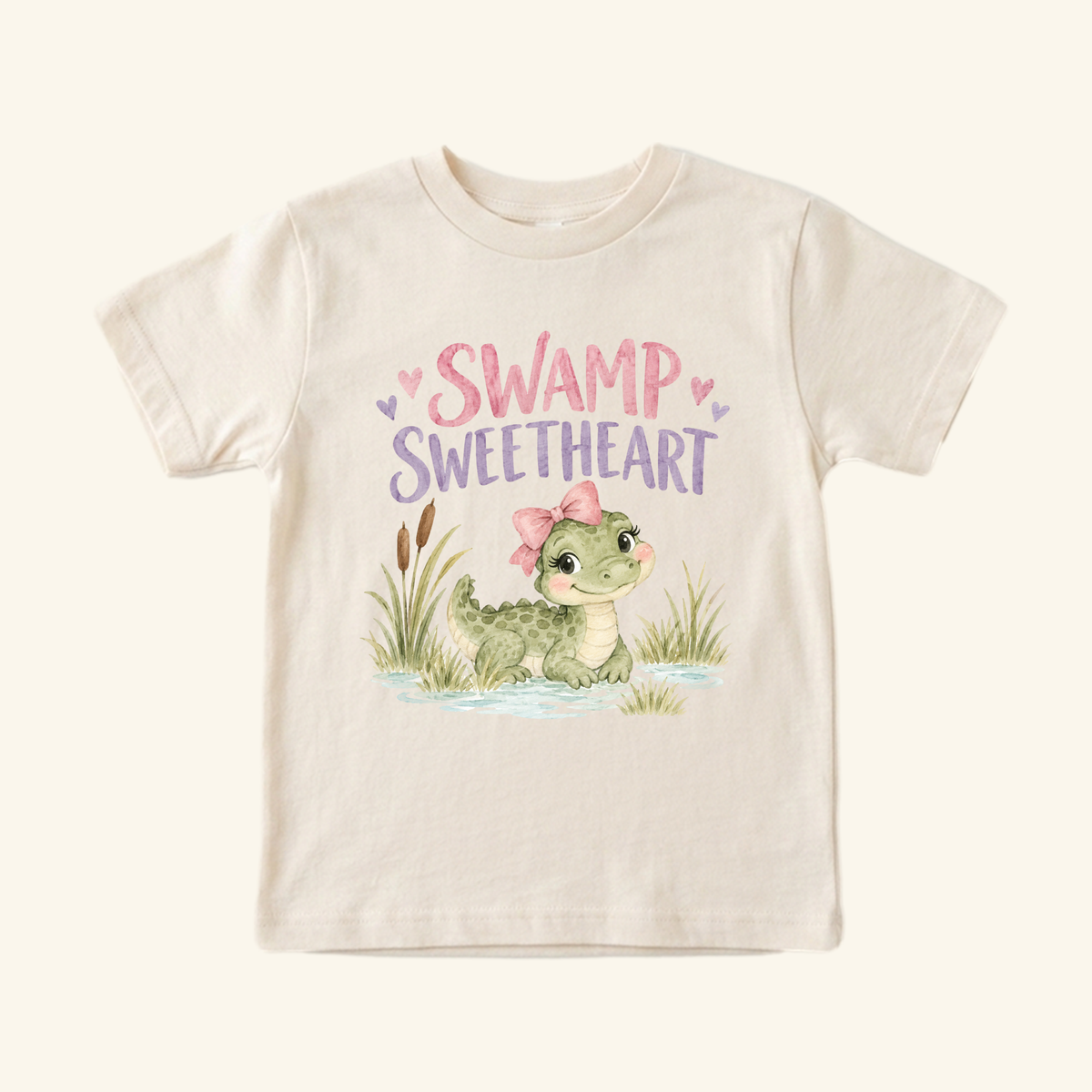 Cream-colored Toodleroos Swamp Sweetheart Toddler Shirt – Baby Alligator Girls Tee featuring a cartoon alligator with a pink bow, surrounded by cattails and the text SWAMP SWEETHEART.