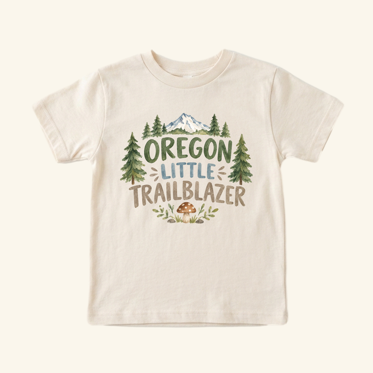 A beige Oregon Toddler Shirt – Little Trailblazer Mountain Forest Tee featuring a mountain and forest graphic with a mushroom illustration and the text Little Trailblazer.
