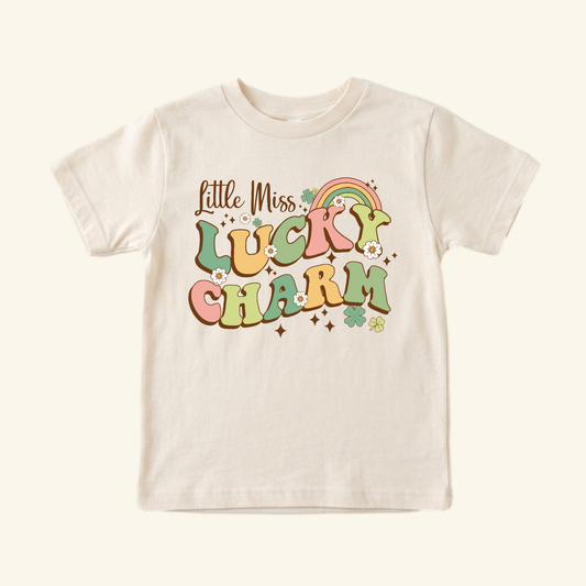 St. Patrick’s Day Toddler Shirt – Little Miss Lucky Charm Tee in cream, featuring retro Little Miss LUCKY CHARM text in pink and green, decorated with a small rainbow, shamrocks, and daisies.