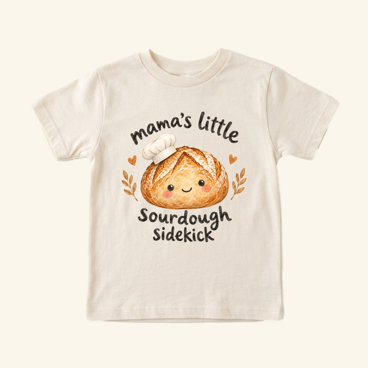 Cream-colored Mama’s Little Sourdough Sidekick Toddler Shirt featuring a smiling loaf of bread wearing a chef’s hat and the text mama’s little SOURDOUGH SIDEKICK.