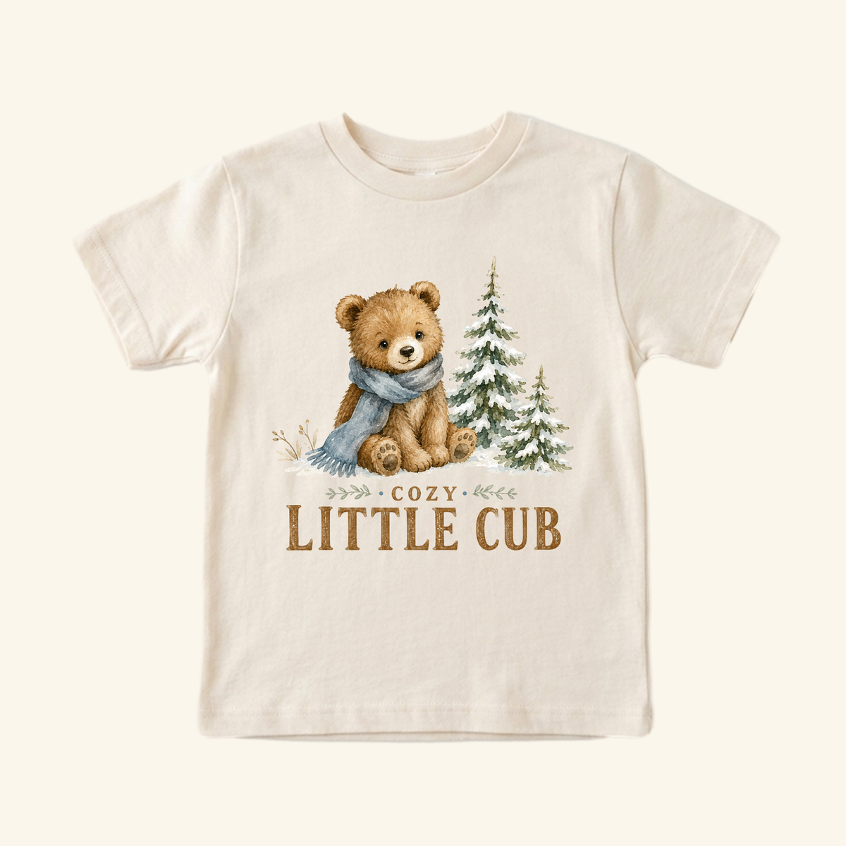 Cream-colored Cozy Little Cub Toddler Shirt featuring a watercolor illustration of a bear cub in a blue scarf sitting next to two pine trees, with the text printed below.