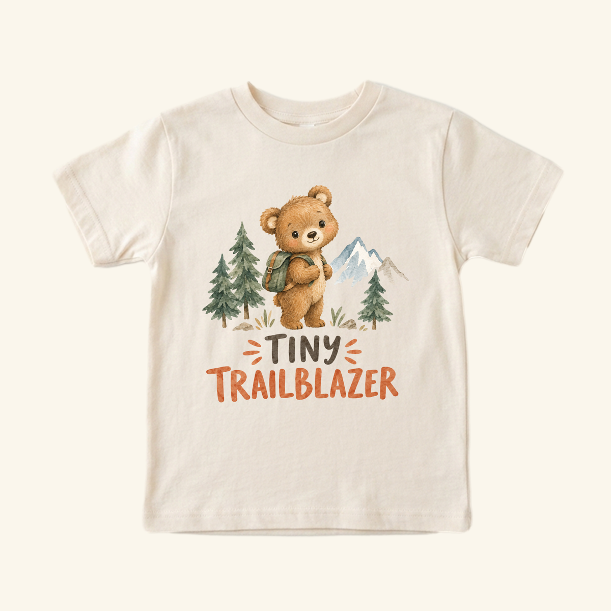 Toodleroos Tiny Trailblazer Toddler Shirt in cream, featuring a watercolor illustration of a bear with a backpack in a forest and the text TINY TRAILBLAZER printed below.