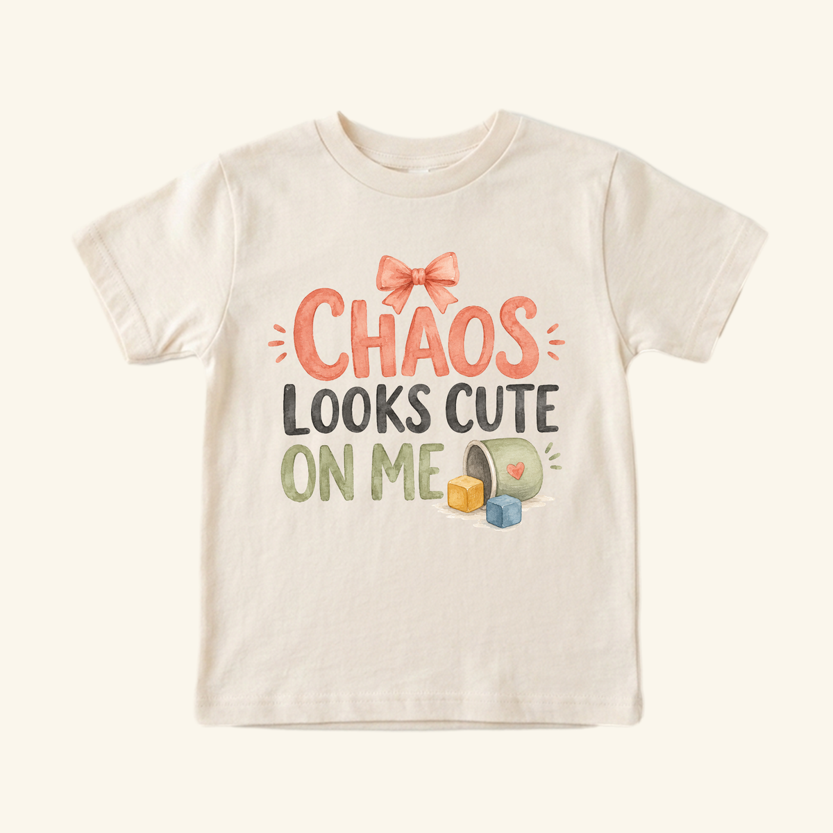 Toodleroos Chaos Looks Cute on Me Toddler Shirt in cream, featuring a bow icon, the text Chaos looks cute on me, and a graphic of a spilled toy bucket and blocks.
