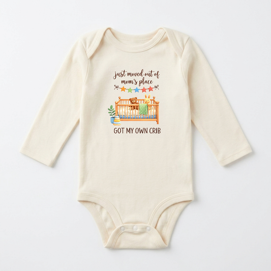 A cream-colored long-sleeve baby bodysuit with a graphic print on the front. The graphic features a cartoon illustration of a bear and rabbit in a crib with a green blanket, accompanied by the text 'just moved out of mom's place GOT MY OWN CRIB' in brown lettering.