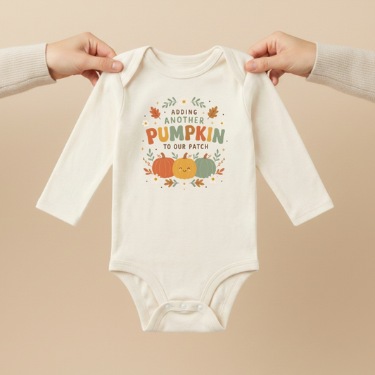 Two hands hold the Toodleroos Long-Sleeve Fall Pregnancy Announcement Onesie – Adding Another Pumpkin, featuring a cream color and charming pumpkin and leaf illustrations with the words “Adding another pumpkin to our patch” on the front, making it the perfect autumn pregnancy reveal outfit.