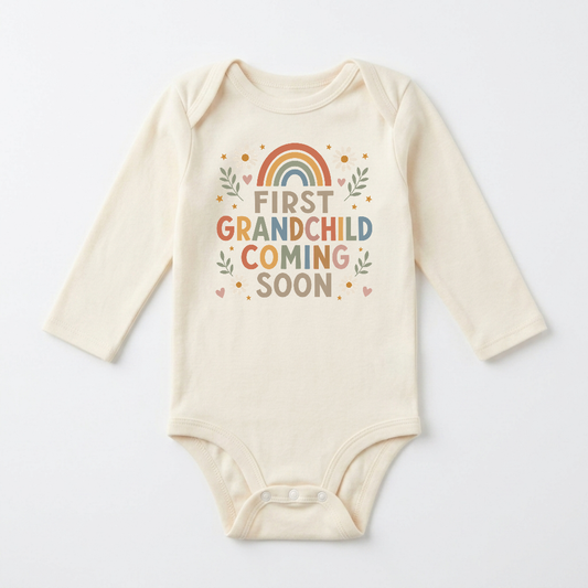 Introducing the Toodleroos Long-Sleeve First Grandchild Pregnancy Announcement Onesie – Coming Soon: a cream-colored onesie decorated with colorful “First Grandchild Coming Soon” lettering, whimsical hearts, leafy accents, and a rainbow motif above the text. This gender-neutral piece is perfect for announcing your first grandchild’s arrival or as a thoughtful pregnancy announcement.