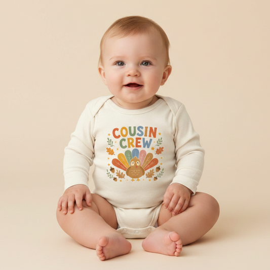 A smiling baby sits on a beige background, wearing the Toodleroos Long-Sleeve Thanksgiving Cousin Baby Onesie – Cousin Crew Turkey Bodysuit, featuring a colorful turkey graphic and “Cousin Crew” on the front—the perfect Thanksgiving outfit for your little one’s cousin crew ensemble.