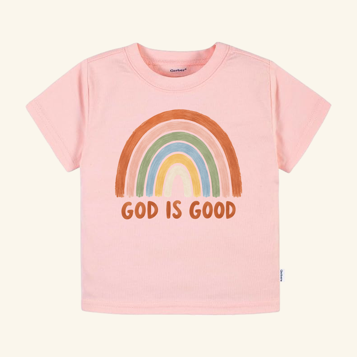 The Toodleroos Christian Toddler Shirt – God Is Good Rainbow Tee features a hand-drawn earthy-toned rainbow and bold orange GOD IS GOOD text on pink.