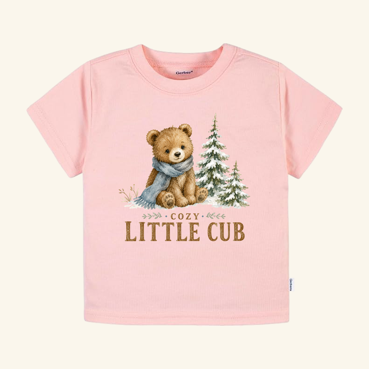 Light pink Cozy Little Cub Toddler Shirt – Cute Winter Bear Tee featuring an illustration of a bear wearing a scarf next to two pine trees, with the woodland winter text printed below.