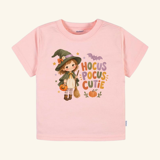 Light pink Toodleroos Hocus Pocus Cutie Witch Toddler Halloween Shirt featuring a cartoon witch holding a pumpkin bucket and broom, with the text Hocus Pocus Cutie and a small bat illustration.