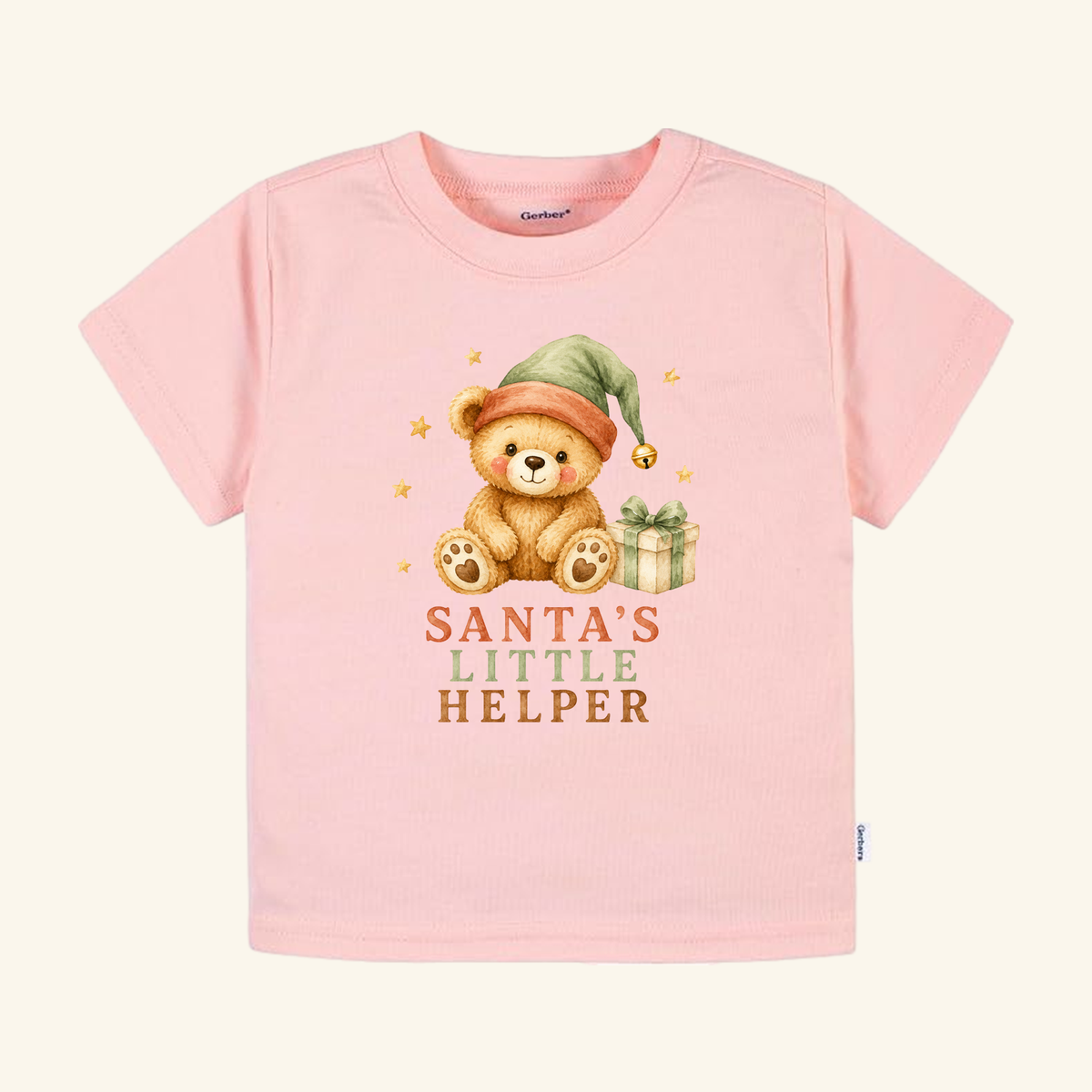 Light pink Toodleroos Santa’s Little Helper Toddler Shirt – Teddy Bear Christmas Tee featuring a teddy bear in a green elf hat next to a gift box, with the text Santa’s Little Helper printed below.