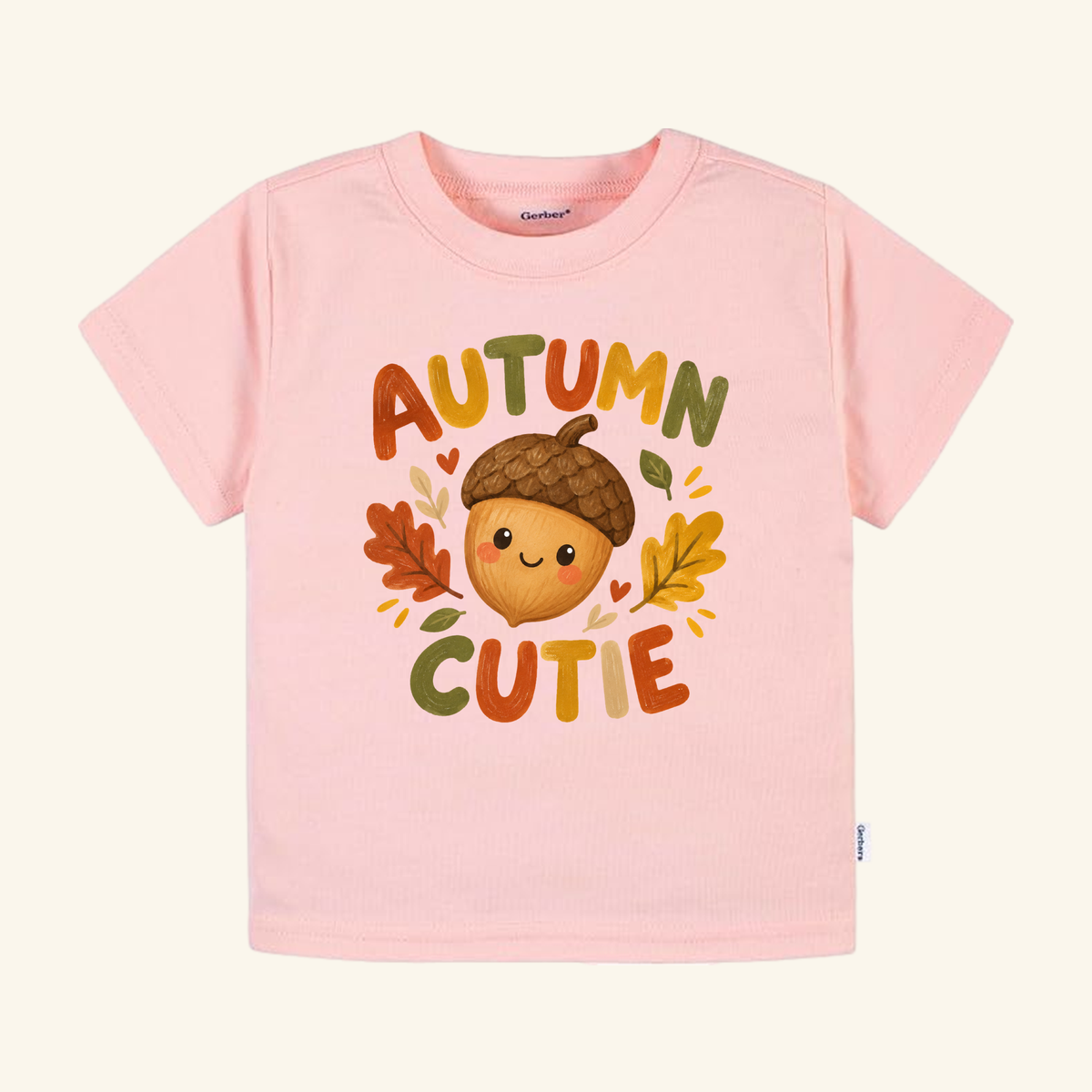 Pale pink Autumn Cutie Acorn Toddler Shirt featuring a graphic of a smiling acorn surrounded by autumn leaves and the text AUTUMN CUTIE.