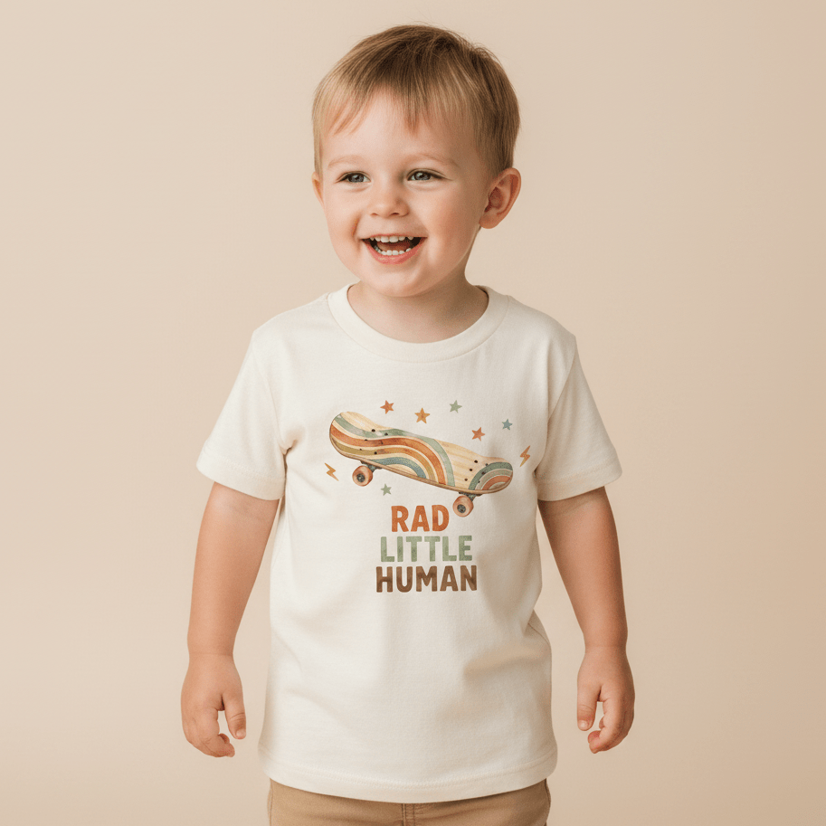 A smiling young child stands against a beige background, wearing the Toodleroos Toddler Skateboard Shirt – Rad Little Human Short-Sleeve Graphic Tee, featuring a cream-colored retro skater style with a rainbow design and “Rad Little Human” printed on the front.