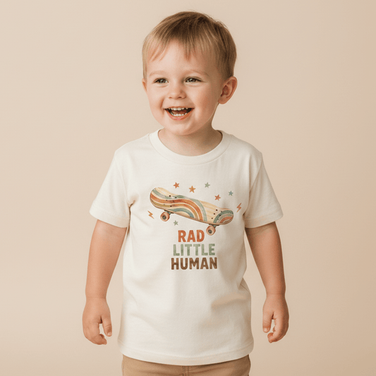 A smiling young child stands against a beige background, wearing the Toodleroos Toddler Skateboard Shirt – Rad Little Human Short-Sleeve Graphic Tee, featuring a cream-colored retro skater style with a rainbow design and “Rad Little Human” printed on the front.