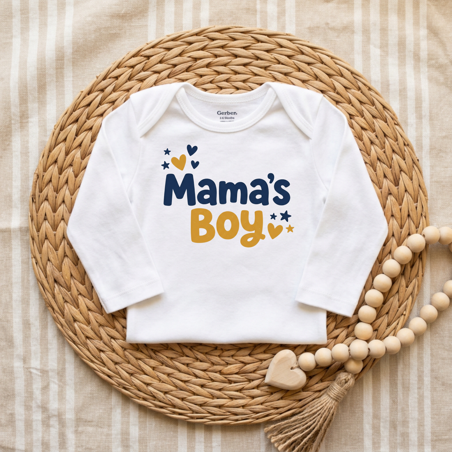 A white long-sleeve baby bodysuit with 'Mama's Boy' printed in navy and mustard yellow, styled flat-lay on a round woven straw mat with a wooden bead garland.