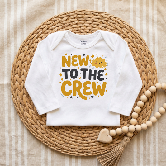 A white long-sleeve baby bodysuit with a colorful 'NEW TO THE CREW' graphic, laid flat on a round woven straw mat with a wooden bead garland.