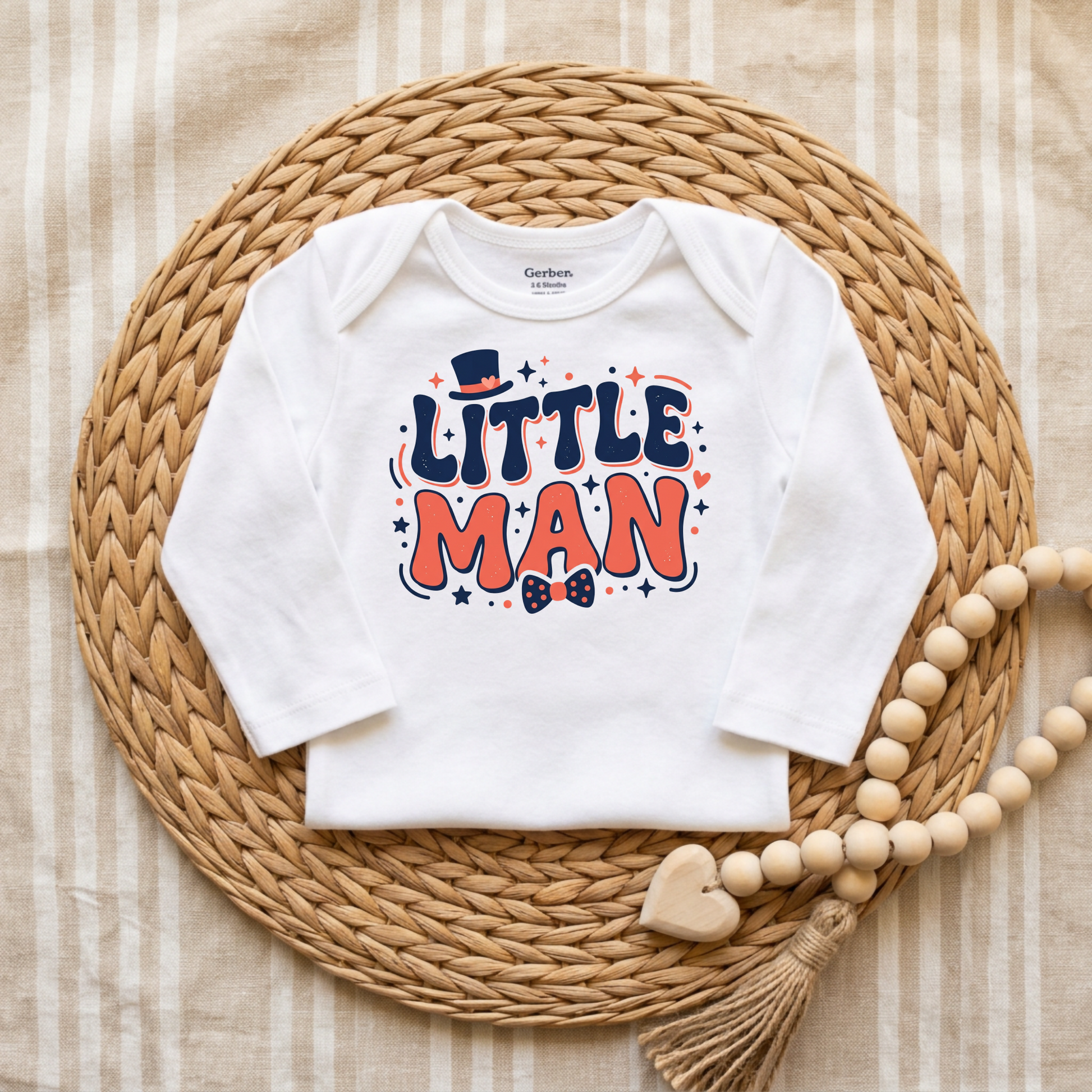 A white long-sleeve baby bodysuit with a colorful 'LITTLE MAN' graphic print, styled flat-lay on a round woven straw mat with a wooden bead garland.