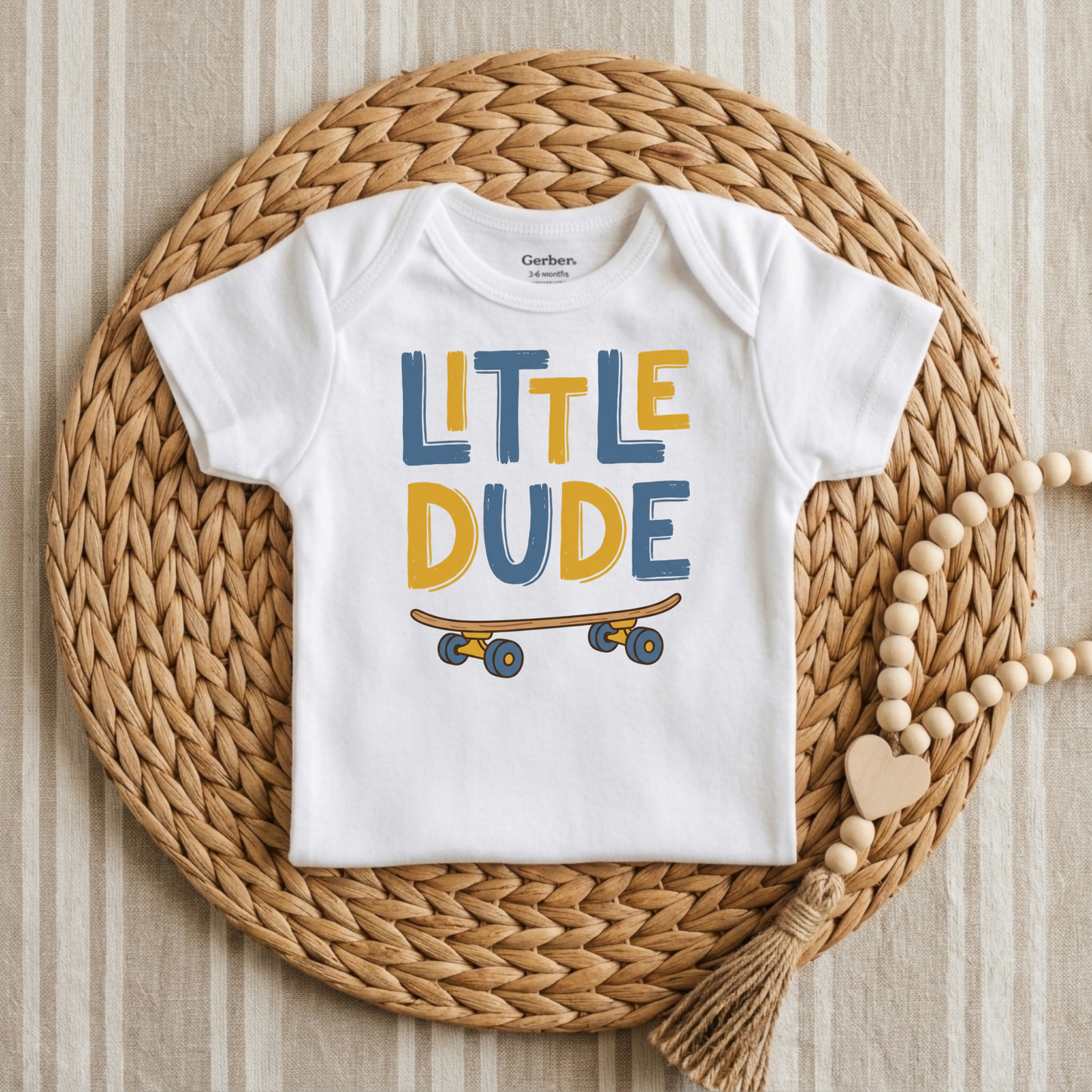 A white short-sleeve baby bodysuit with 'LITTLE DUDE' printed in blue and yellow letters above a skateboard graphic, laid flat on a woven round mat with a wooden bead necklace nearby.