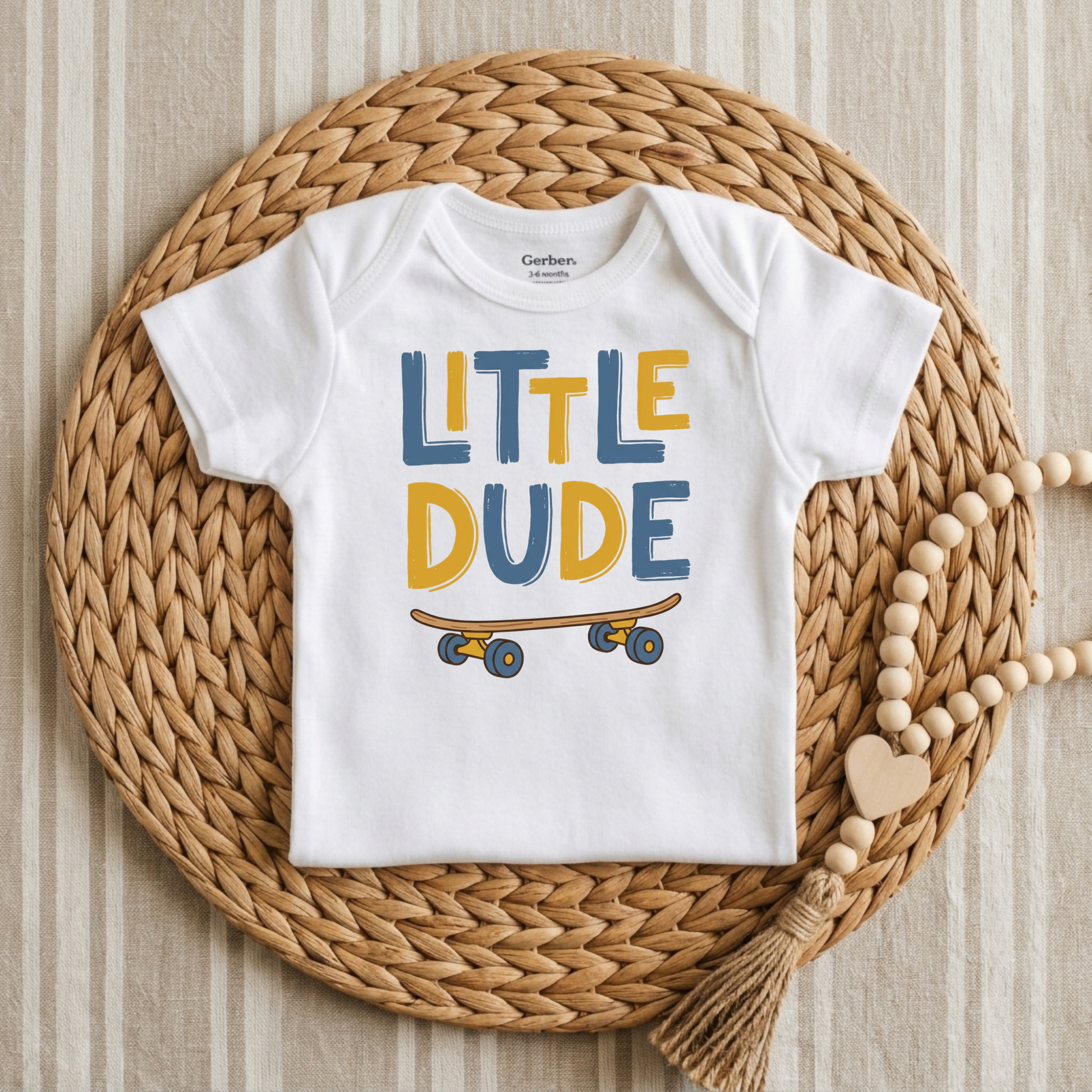 A white short-sleeve baby bodysuit with 'LITTLE DUDE' printed in blue and yellow letters above a skateboard graphic, laid flat on a woven round mat with a wooden bead necklace nearby.