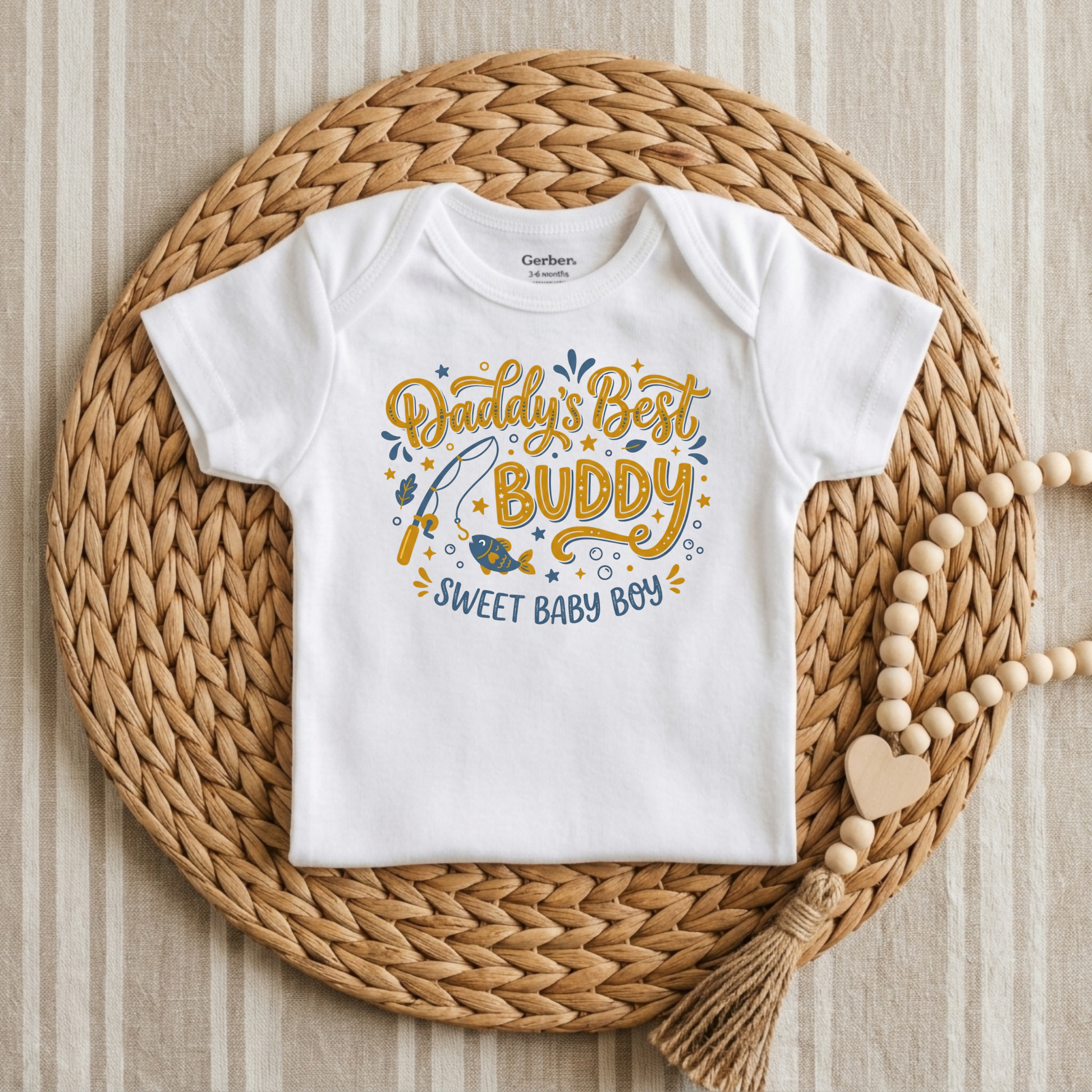 A white short-sleeve baby bodysuit laid flat on a round woven straw mat, with a decorative wooden bead strand nearby. The bodysuit has a colorful front graphic reading “Daddy’s Best Buddy Sweet Baby Boy” with fish and star details.