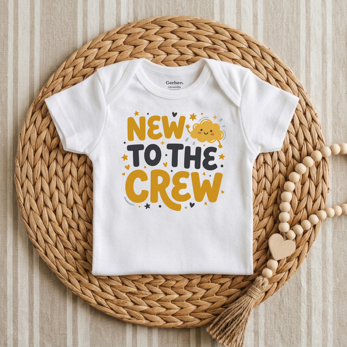 A white short-sleeve baby bodysuit with a colorful graphic reading 'NEW TO THE CREW' in yellow, black, and gray, styled flat on a round woven straw mat with a wooden bead tassel.