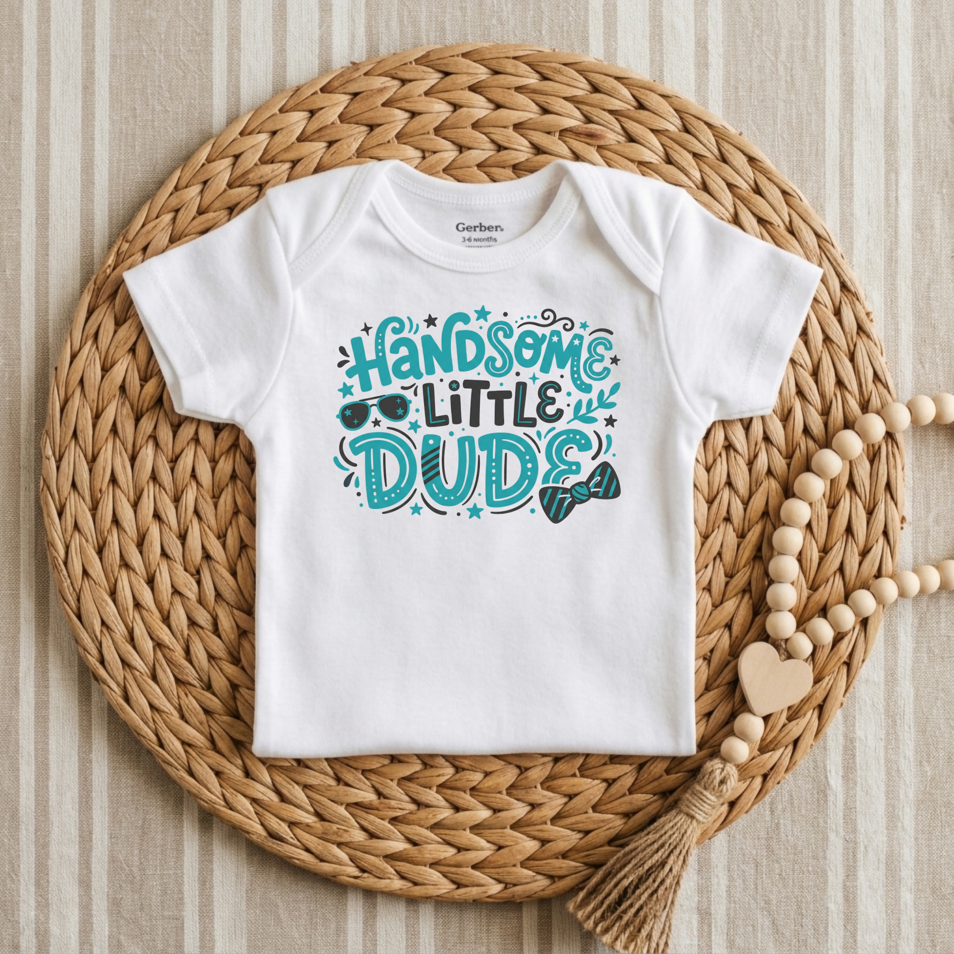 A white short-sleeve baby bodysuit with a colorful graphic reading 'HANDOME LITTLE DUDIE' in teal and blue, styled flat on a round woven straw mat with a wooden bead necklace nearby.