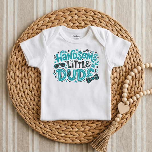 A white short-sleeve baby bodysuit with a colorful graphic reading 'HANDOME LITTLE DUDIE' in teal and blue, styled flat on a round woven straw mat with a wooden bead necklace nearby.