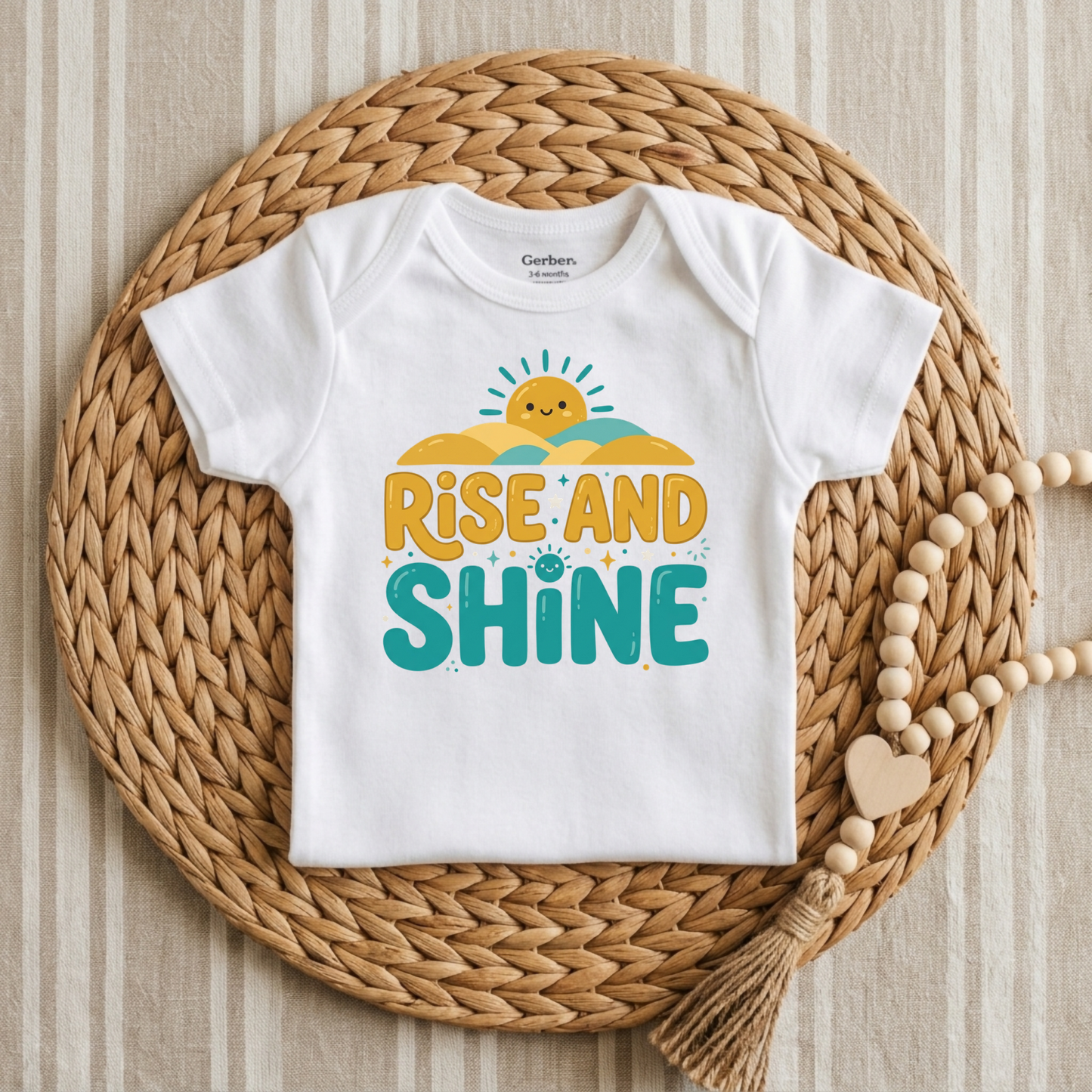 A white short-sleeve baby bodysuit laid flat on a round woven straw mat, with a colorful 'Rise and Shine' graphic on the front. The bodysuit has a visible Garterbaby neck label.