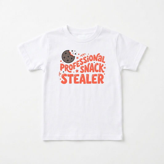 A white short-sleeve kids' t-shirt with a graphic on the chest that reads 'PROFESSIONAL SNACK STEALER' in orange-red lettering, accompanied by a small chocolate chip cookie illustration.