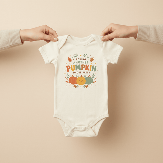 Two hands hold up the Toodleroos Fall Pregnancy Announcement Onesie – Adding Another Pumpkin Short-Sleeve Bodysuit, which features colorful pumpkins and leaves on a white background—a perfect baby announcement for a Fall pregnancy. The text on the bodysuit reads, “Adding another pumpkin to our patch.”.
