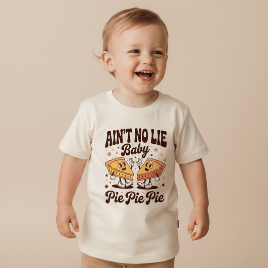 A cheerful toddler sports the Toodleroos Funny Toddler Thanksgiving Shirt – Ain’t No Lie Baby Pie Pie Pie Tee, featuring two pie slices and playful “Ain’t No Lie Baby, Pie Pie Pie” text—a perfect humorous Thanksgiving outfit for your little one.