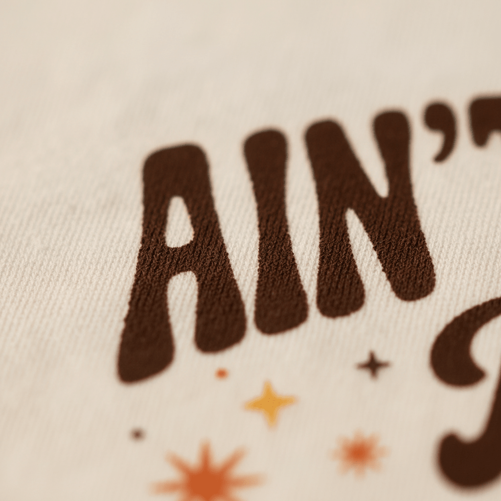 Close-up of textured fabric featuring the brown text “AIN” and small orange and yellow star illustrations on a light background, from the Toodleroos Funny Toddler Thanksgiving Shirt – Ain’t No Lie Baby Pie Pie Pie Tee, perfect for a pie graphic tee or a humorous Toddler Thanksgiving outfit.