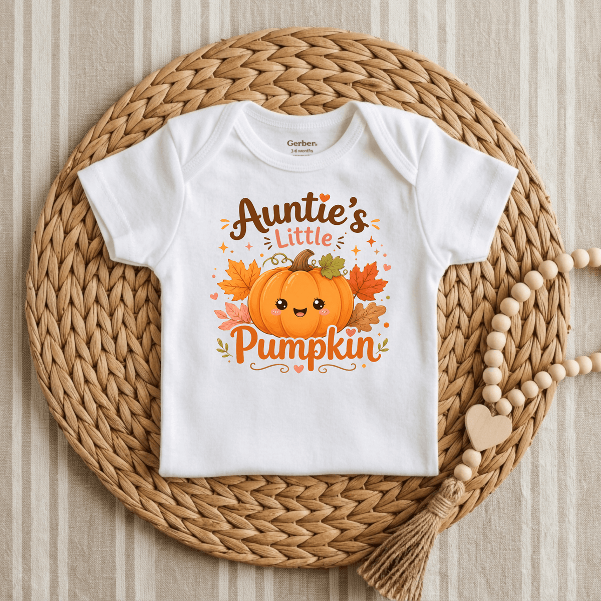 Showcased on a round woven mat next to a wooden beaded garland with a heart and tassel, the Toodleroos Auntie’s Little Pumpkin Baby Onesie – Fall Pumpkin Bodysuit features the phrase “Aunties Little Pumpkin” alongside an adorable pumpkin with leaves. This charming autumn baby bodysuit is the perfect pumpkin patch outfit for your little one.