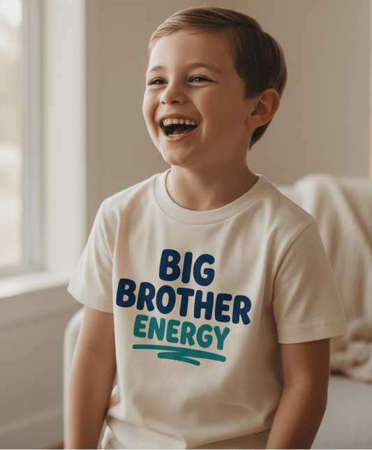 A cheerful young boy wears the Toodleroos Big Brother Energy Toddler Shirt – Confident Sibling Tee, featuring bold blue and teal lettering, as he proudly sits indoors near a window after his exciting promotion to big brother.