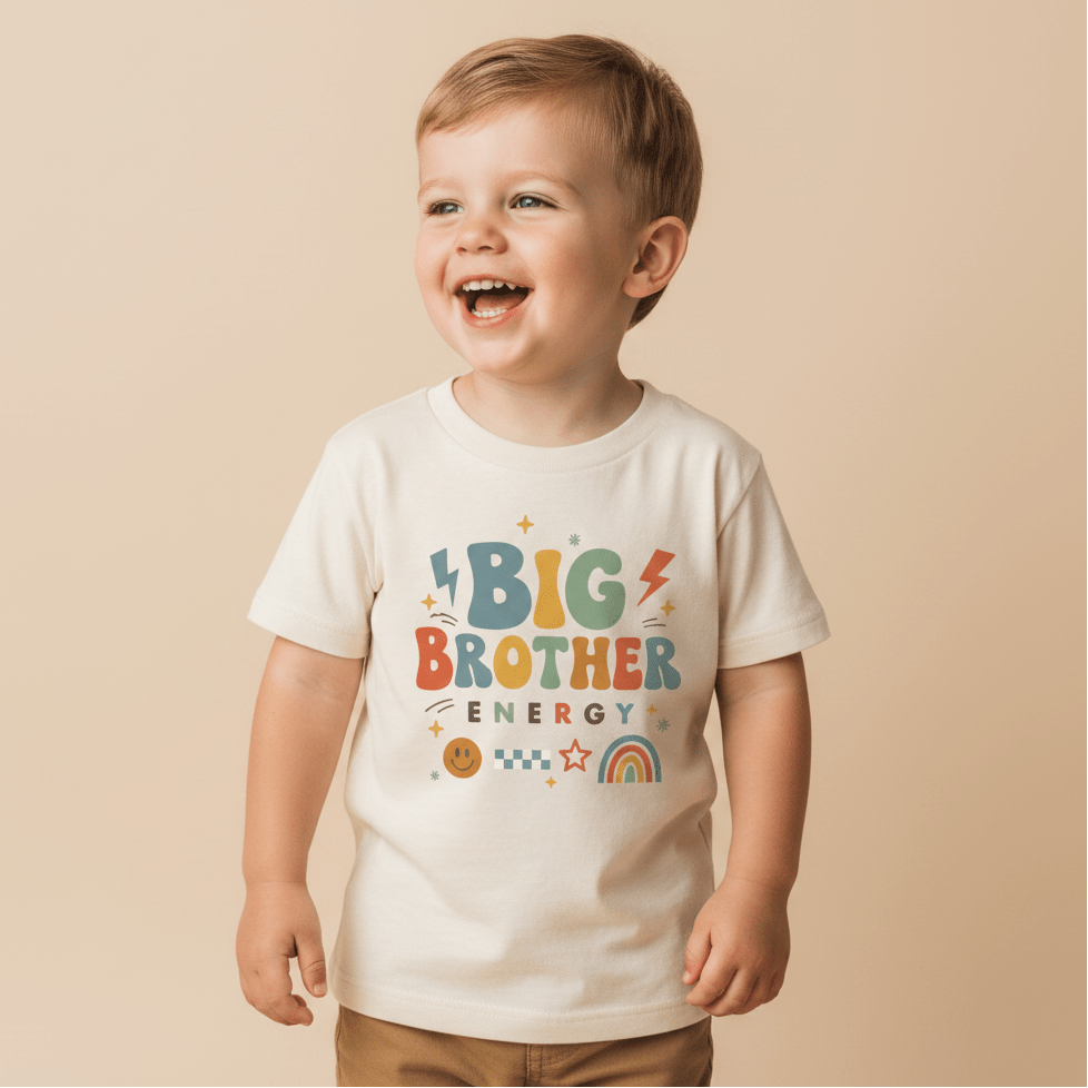 A smiling young child stands against a beige background, wearing the Toodleroos Big Brother Energy Toddler Shirt – Retro Graphic Design, featuring colorful text and playful symbols like a rainbow, smiley face, and lightning bolts—perfect for a fun pregnancy announcement.
