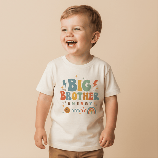 A smiling young child stands against a beige background, wearing the Toodleroos Big Brother Energy Toddler Shirt – Retro Graphic Design, featuring colorful text and playful symbols like a rainbow, smiley face, and lightning bolts—perfect for a fun pregnancy announcement.