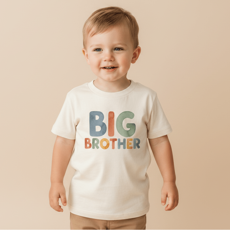A smiling young child wears the Toodleroos Big Brother Toddler Shirt – Colorful Retro Design, featuring bold and vibrant “BIG BROTHER” lettering on a cream-colored shirt. Ideal for pregnancy announcements, the scene is set against a solid beige background.