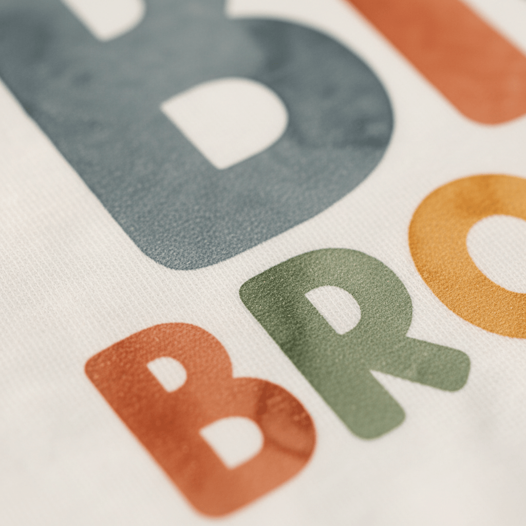 Close-up of the Big Brother Toddler Shirt – Colorful Retro Design by Toodleroos, featuring vibrant block letters that spell ‘BRO’ in blue, green, and orange on a white fabric background. The visible texture of both the fabric and ink adds charm to this playful toddler shirt—an ideal choice for a cheerful pregnancy announcement or as a cute big brother reveal.