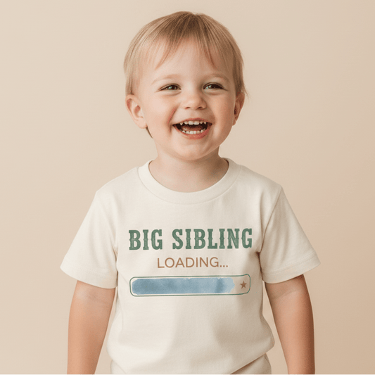 A happy young child models the Toodleroos Big Sibling Loading Toddler Shirt – Pregnancy Announcement, featuring “BIG SIBLING LOADING…” and a partially filled loading bar on a white t-shirt, set against a neutral backdrop. This charming shirt is the perfect way to share your growing family’s news!.