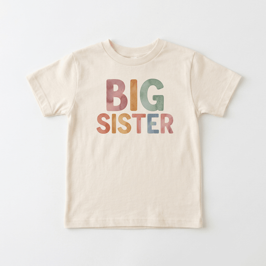 The Toodleroos Big Sister Toddler Shirt – Colorful Retro Design features a cream-colored fabric with “BIG SISTER” boldly printed in large, pastel, retro-inspired letters across the front—perfect for toddlers or as an adorable pregnancy announcement—shown here on a plain white background.