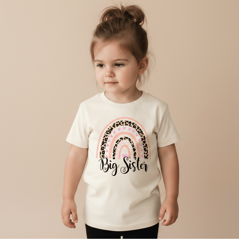 A young child with brown hair in a ponytail wears the Toodleroos Big Sister Toddler Shirt – Leopard Rainbow Design, which features a white base, a pink and leopard print rainbow, and “Big Sister” on the front. She stands against a plain beige background, looking slightly to the side—perfect for a sweet pregnancy announcement.