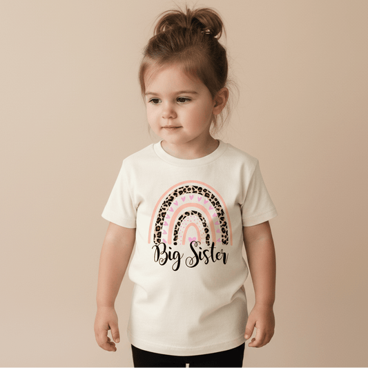 A young child with brown hair in a ponytail wears the Toodleroos Big Sister Toddler Shirt – Leopard Rainbow Design, which features a white base, a pink and leopard print rainbow, and “Big Sister” on the front. She stands against a plain beige background, looking slightly to the side—perfect for a sweet pregnancy announcement.