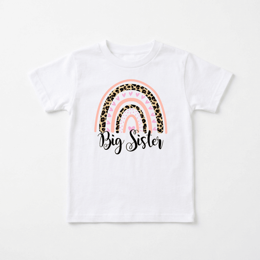 The Big Sister Toddler Shirt – Leopard Rainbow Design by Toodleroos features a white tee adorned with a pink, gold, and leopard print rainbow. The phrase “Big Sister” is written in elegant black cursive beneath the rainbow, creating an adorable top that’s perfect for big sister announcements or pregnancy reveals.