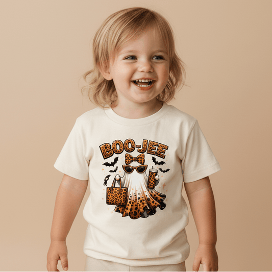 A happy toddler models the Toodleroos Boo-Jee Ghost Toddler Girls Shirt—a leopard print Halloween tee featuring a ghost with sunglasses—styled with a matching leopard-print bow and bag, festive pumpkins, and bats in the background.