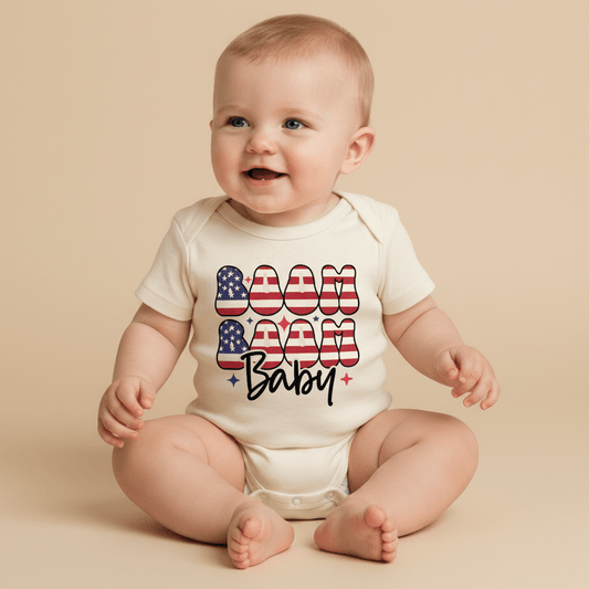 A happy baby sits on a beige background, dressed in the Toodleroos Boom Boom Baby 4th of July Onesie – Patriotic Bodysuit, a gender-neutral white outfit featuring “Boom Boom Baby” in bold American flag lettering—an ideal choice for your little one’s Independence Day celebration.