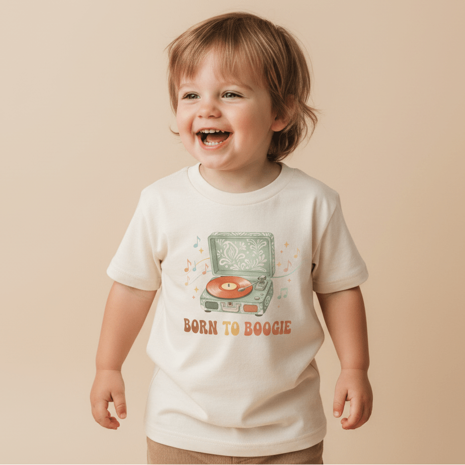 A smiling young child wears the Toodleroos Toddler Short-Sleeve Music Shirt – Born to Boogie Retro Record Player Tee, featuring an illustration of a retro record player, colorful musical notes, and “BORN TO BOOGIE” text on the front, standing against a beige background.