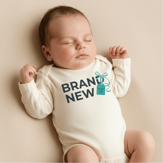 A sleeping baby in a cream-colored Toodleroos “Brand New Baby Onesie – Welcome Little One Bodysuit” that says BRAND NEW, with a small illustrated gift tag beside the text—perfect as a baby shower gift or as a sweet baby announcement outfit.